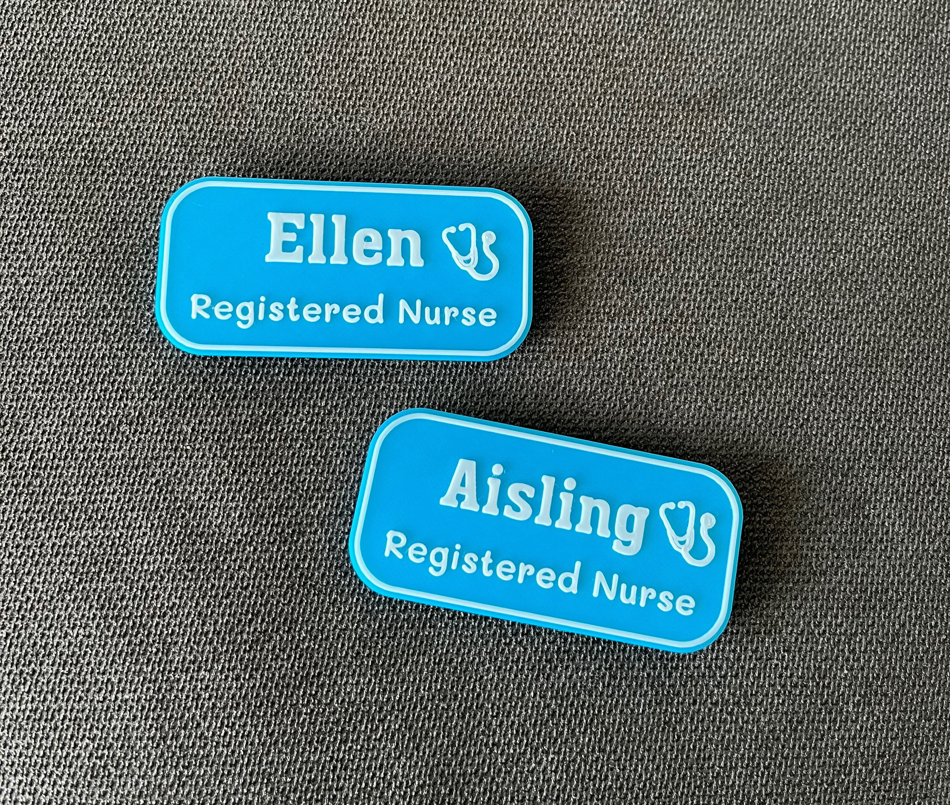 Two matching blue Kind Badges for Ellen and Aisling, both Registered Nurses, displayed on dark fabric showing professional coordination